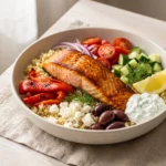 Mediterranean salmon bowl with roasted salmon, lemon quinoa, cherry tomatoes, cucumber, olives, feta and cucumber tzatziki in a white ceramic bowl