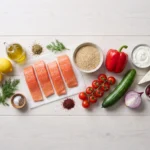 Raw ingredients laid out on a white wood surface: four salmon fillets, dry quinoa, red bell pepper, cucumber, cherry tomatoes, Greek yogurt, feta, Kalamata olives, lemons, dill and olive oil