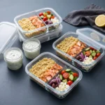 Glass meal prep containers holding quinoa, flaked salmon, cucumber and tomato salad, red onion, olives and feta, with tzatziki in separate jars