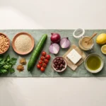 Raw ingredients for a chickpea bowl: chickpeas, dry quinoa, cucumber, tomatoes, feta, olives, tahini and lemons