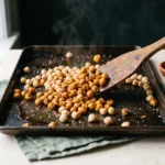 Half-roasted chickpeas being tossed with a wooden spatula on a hot bare sheet pan