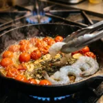 Raw shrimp being laid with tongs into a skillet of blistered cherry tomatoes and sliced garlic over a gas flame