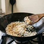 Pale cauliflower grains dry-sauteing in a carbon-steel wok, a third of them freckled golden, spatula caught mid-turn.