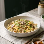 A bowl of cauliflower fried rice with golden-freckled grains, soft egg ribbons, mushrooms and sliced scallion greens.