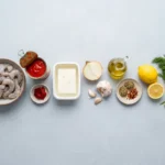Raw ingredients for garides saganaki laid out overhead: peeled shrimp, canned tomatoes, tomato paste, block feta, onion, garlic, olive oil, oregano, lemon and dill