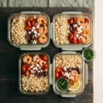 Four glass meal-prep containers of Mediterranean shrimp and cauliflower rice with tomatoes, olives and feta