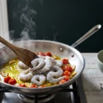 Still-frozen grey shrimp added in a single layer to a skillet of blistered cherry tomatoes and sliced garlic