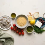 Raw shrimp, dry orzo, feta, olives, lemon, tomatoes and dill laid out on a pale worktop
