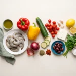 Raw shrimp, whole bell peppers, zucchini, red onion, cherry tomatoes, feta, olives and lemon laid out on a pale worktop