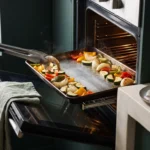 A half-sheet pan of part-roasted peppers and zucchini pulled from the oven, tongs clearing a space for the shrimp