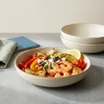 One serving of roasted shrimp, peppers, zucchini and tomatoes in a shallow stoneware bowl with feta and a lemon wedge