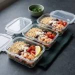 Three glass meal-prep containers packed with shrimp couscous, olives, feta and cherry tomatoes on a dark slate worktop