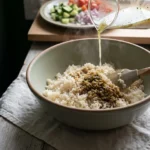 Lemon-oregano dressing poured over warm cooked rice in a wide bowl, a spatula folding one edge