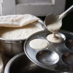 Risen fermented batter being ladled into a greased stainless steel idli mould plate