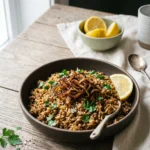 Bowl of vegan mujadara with brown rice topped with dark crispy onions, parsley and a lemon wedge