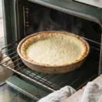 Cheese crust part-baked in a pie dish, fused into a pale sheet with the rim just turning gold, no filling in it