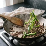 Sliced green pepper, onion and mushrooms cooking with browned ground beef in a wide carbon steel skillet