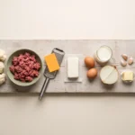 Raw cauliflower, ground beef, cheddar, cream cheese, eggs and cream laid out on a wooden board