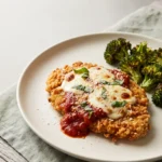 Keto chicken parmesan meal prep cutlet with a golden pork rind crust, melted mozzarella and roasted broccoli on a cream plate