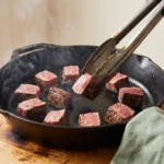 Sirloin cubes searing in a single uncrowded layer in a hot cast iron skillet, tongs turning one cube