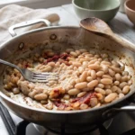 A fork crushing a third of the cannellini beans in a pan with softened onion and darkened tomato paste