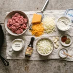 Raw ingredients for a keto ground beef casserole laid out overhead, including beef, cheddar, mozzarella, and spices