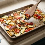 Roasted zucchini, pepper, onion, and chickpeas mid-toss on a sheet pan for a Mediterranean couscous bowl