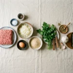 Raw ground beef, grated onion, egg, breadcrumbs, parsley, oregano, garlic and olive oil laid out for keftedes.